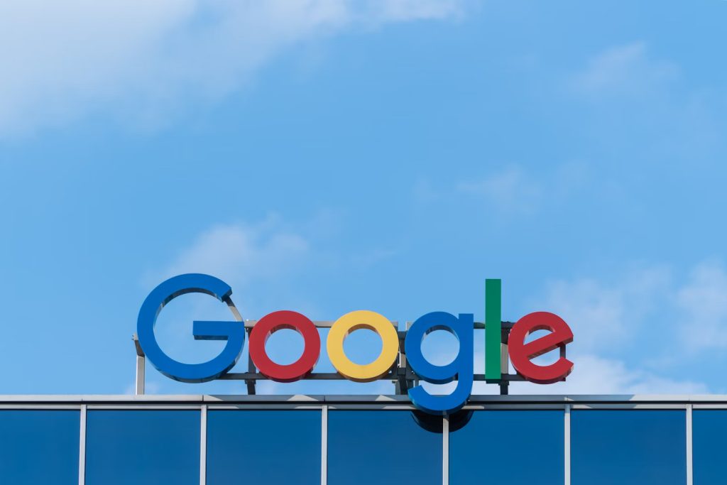: A large, colorful Google logo sign with its distinct red, yellow, blue, and green letters mounted on top of a modern building with blue reflective windows, set against a bright blue sky with scattered clouds.