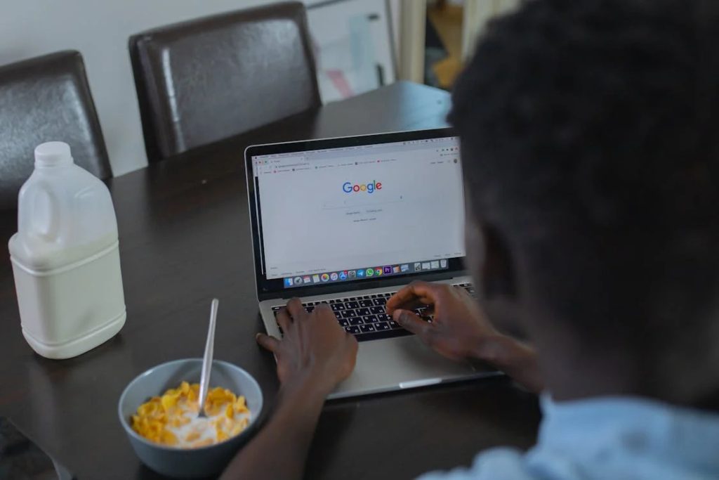 A person viewing the Google search homepage on a laptop while eating cereal.