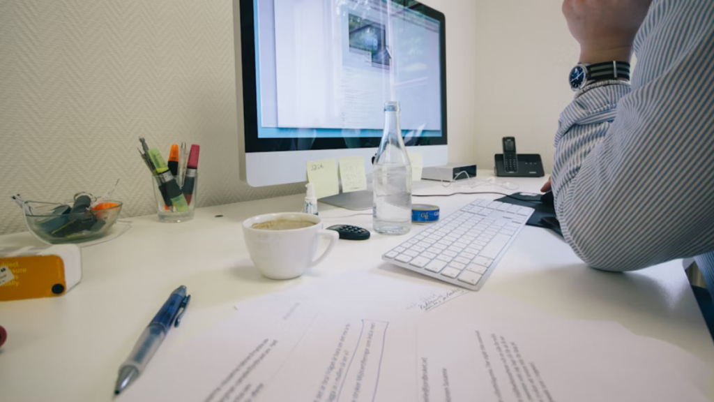 A person working on a computer at a desk with papers, a coffee cup, and a pen, illustrating the work involved when considering what is an example of a whitepaper.