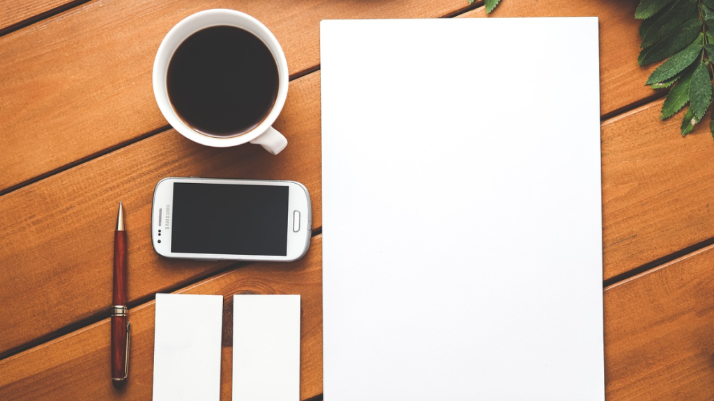 A top-down view of a desk with a coffee cup, smartphone, pen, two business cards, and a blank sheet of paper, representing the tools used to create content like what is an example of a whitepaper.