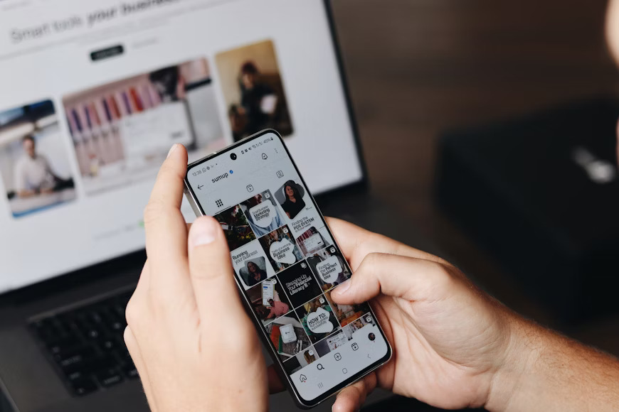 Close-up of hands scrolling through an Instagram feed on a smartphone, with a laptop in the blurred background.