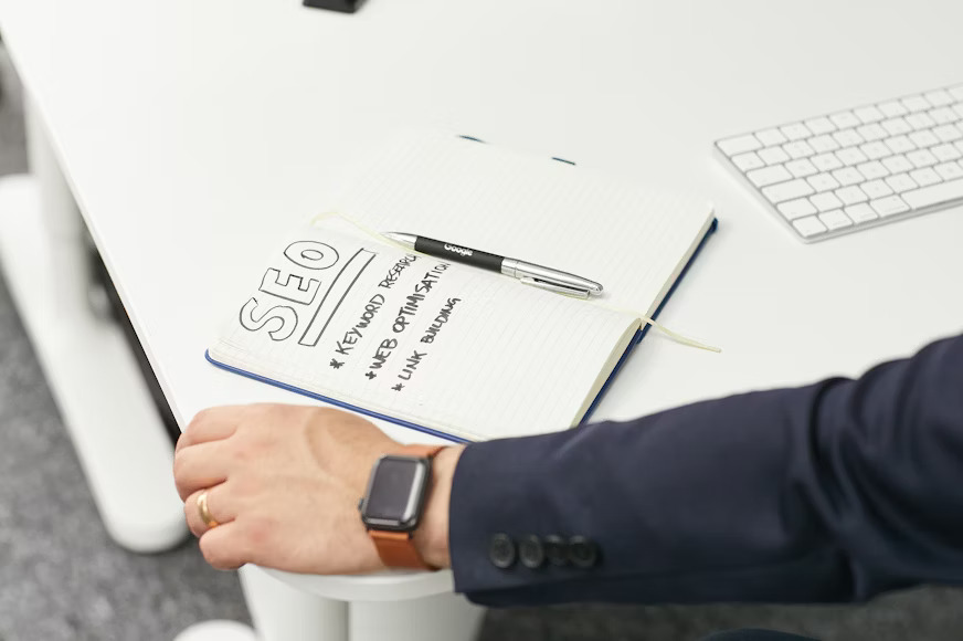 A notebook open to a page with "SEO" written largely, followed by bullet points: Keyword Research, Web Optimization, Link Building, with a pen resting on it.